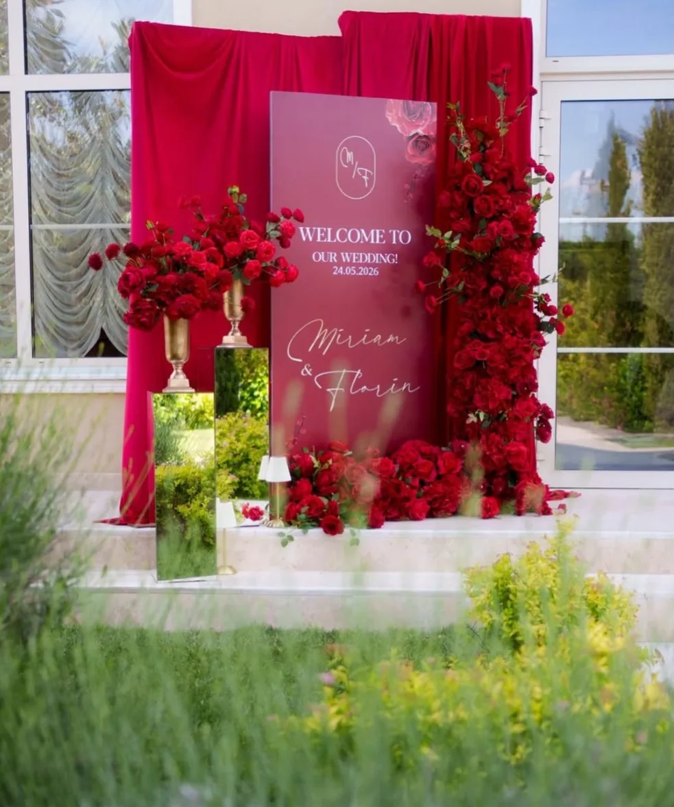 Panou de bun venit bordo cu numele mirilor, încadrat de trandafiri roșii, draperie de catifea și vase aurii